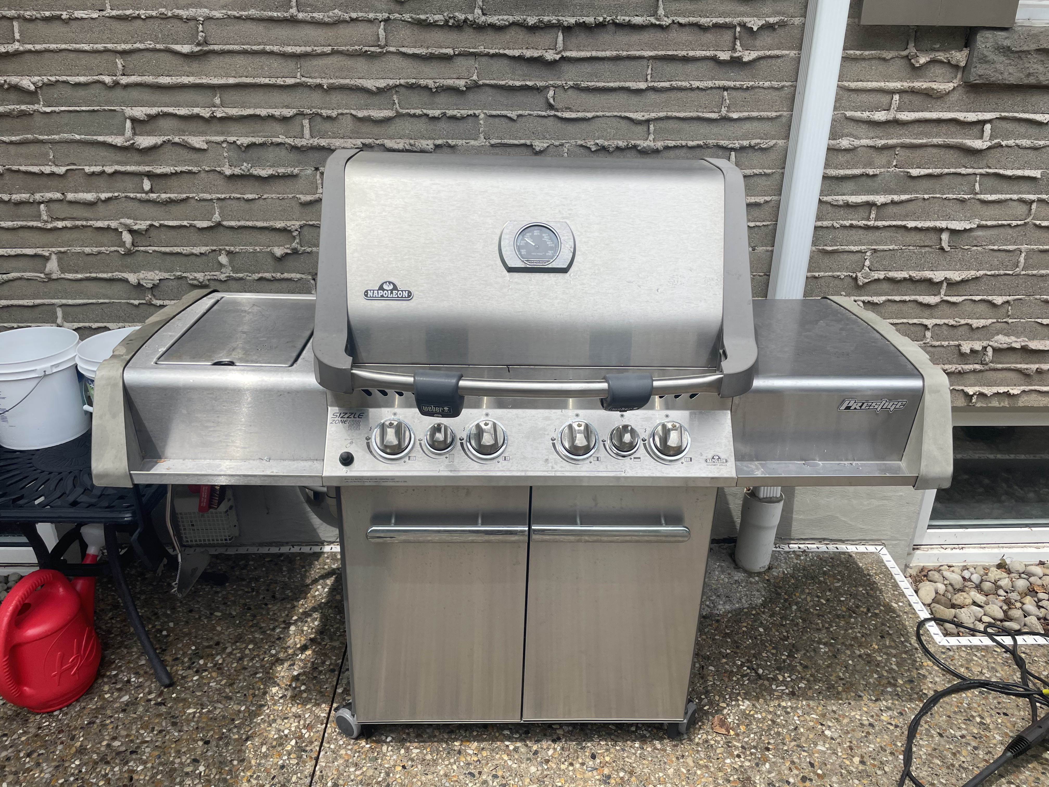 Napoleon Prestige gas grill closed against a brick wall before an Aspect deep clean, its stainless lid and cart dulled by grease film and grime